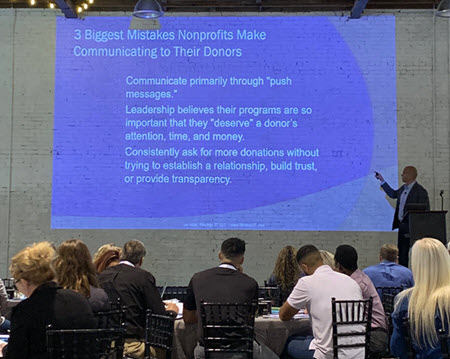 Presentation screen and audience with text: "3 biggest mistakes nonprofits make communicating to their donors: communicate primarily through "push messages," leadership believes their programs are so important that they "deserve" a donor's attention, time and money; consistently ask for more donations without trying to establish a relationship, build trust, or provide transparency"