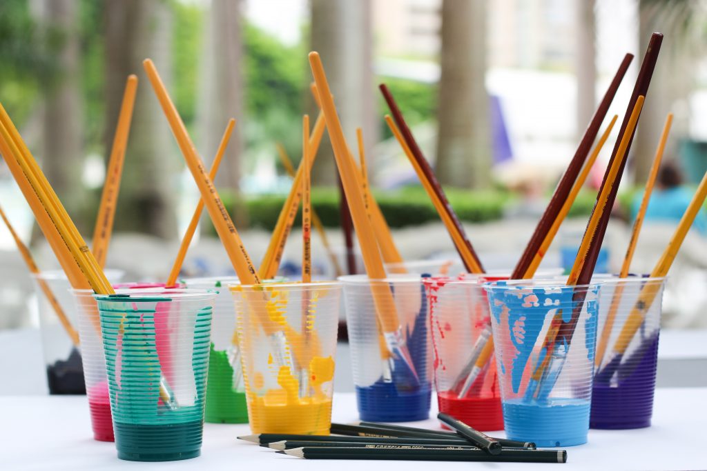 Paint brushes in clear, plastic cups full of paint.