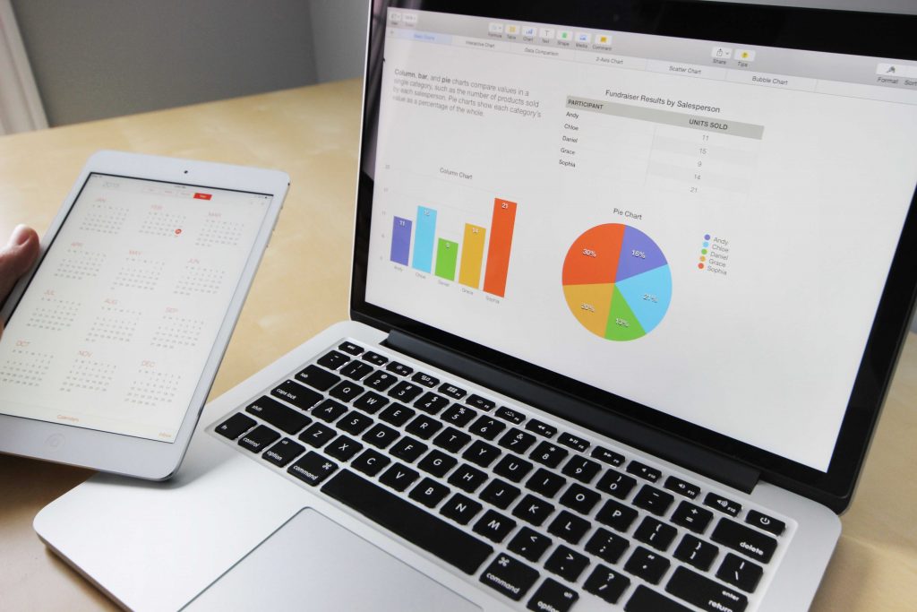 Image of a bar graph and a pie graph on a laptop. A person's hand is visible next to the laptop holding a tablet with a calendar app on the screen.