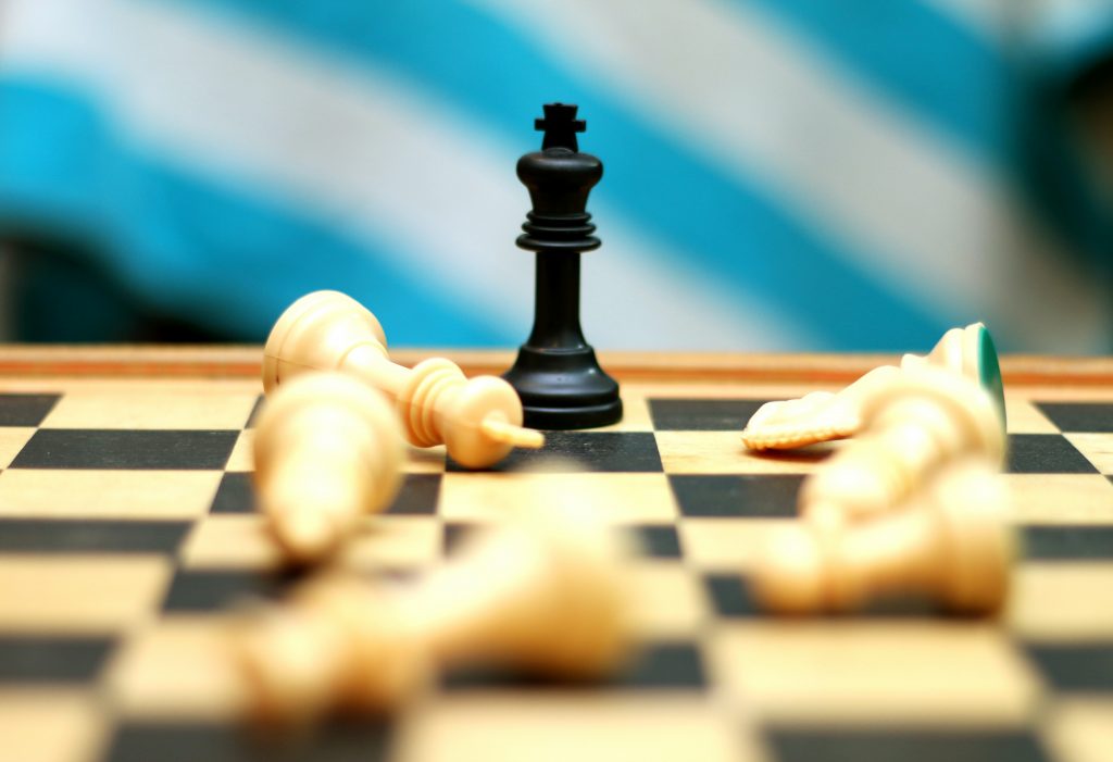 Chessboard with black king piece standing surrounded by fallen white chess pieces.