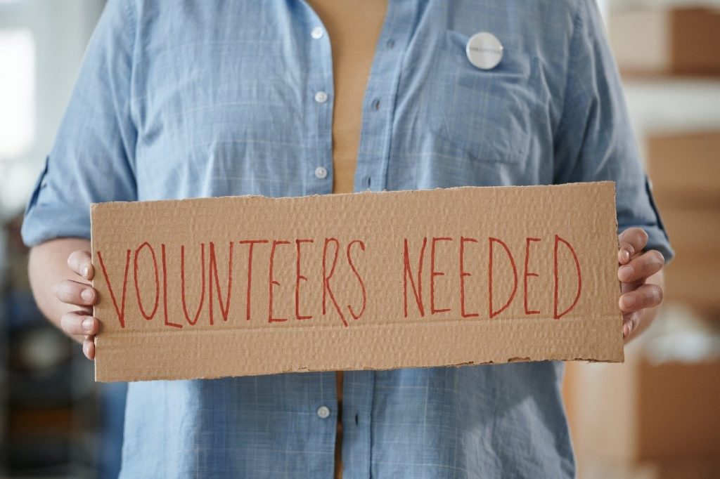 Woman holding sign that says Volunteers Needed
