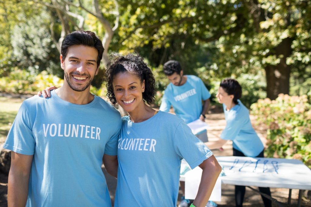 A man and woman wearing blue volunteer shirts looking at the camera and smiling. Two more volunteers work in the background.