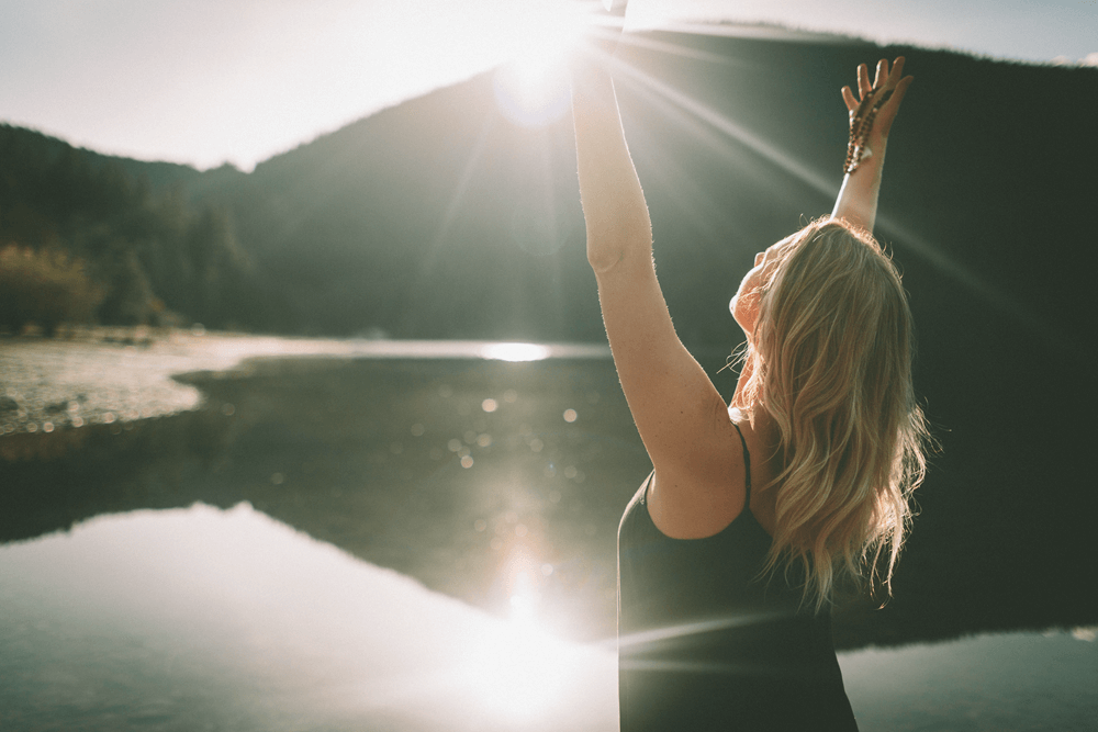 Woman standing next to water with her arms raised, practicing self care