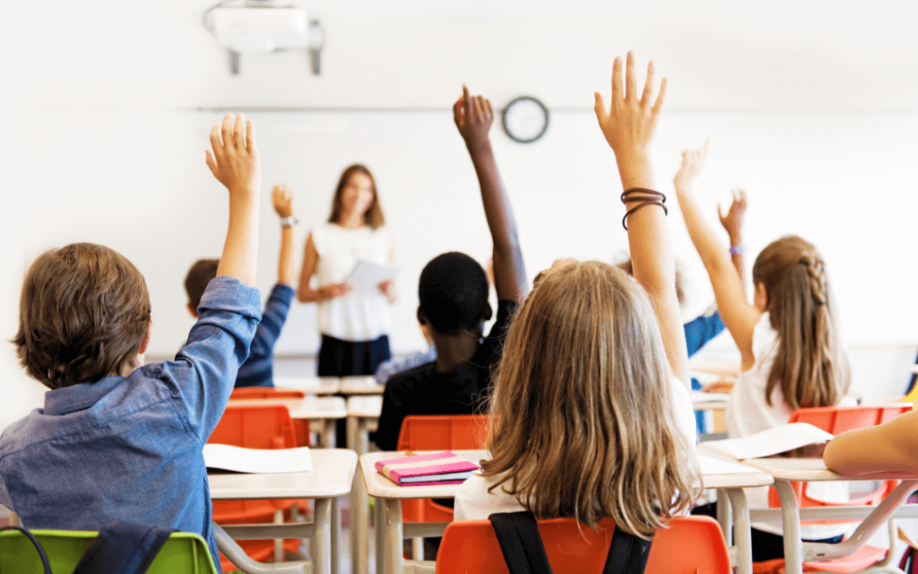 Students in class with their hands up and teacher in the background