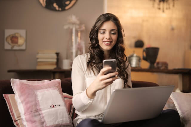 A woman sits in front of an open laptop with her cell phone in her hand.