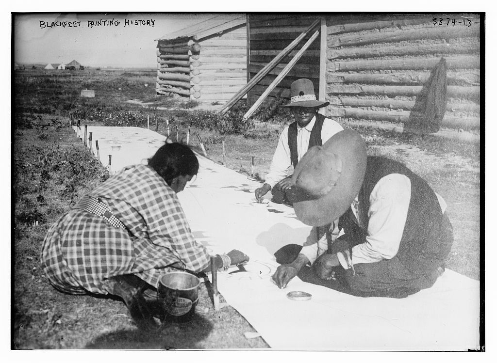 Photograph shows Blackfeet (Sihasapa) Indians painting on a hide next to a log building.